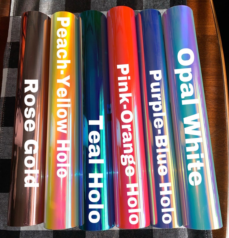 May include: A collection of vinyl rolls in various colors, including rose gold, peach-yellow holo, teal holo, pink-orange holo, purple-blue holo, and opal white. Each roll has a glossy finish and is labeled with its color name in white text.