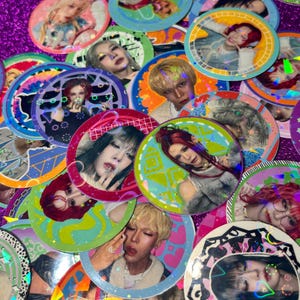 May include: A collection of colorful, circular stickers featuring portraits of various people with diverse hairstyles and makeup. The stickers have holographic and glittery accents, with a purple background.