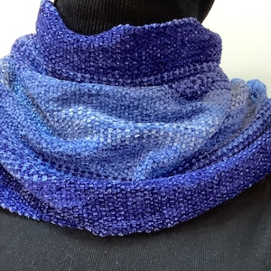 May include: A blue and purple knit cowl scarf. The scarf is a blend of dark and light blues, creating a gradient effect. The fabric has a textured, nubby appearance, and is worn around the neck.