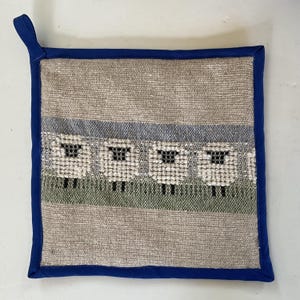 May include: Square beige and blue pot holder with a woven design featuring four white sheep with black faces and legs. The pot holder has a blue border and a loop for hanging.
