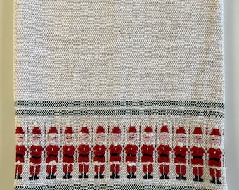 Puede incluir: Un paño de cocina beige con temática navideña con una textura tejida. El borde inferior presenta una fila de figuras de Papá Noel bordadas en rojo y negro, con rayas verdes arriba y abajo.
