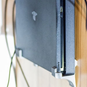 May include: A black PlayStation 4 console mounted on a wall with a white bracket. The console is connected to a power cord and has a USB port visible. The text "PS4" and "SONY" are visible on the console.