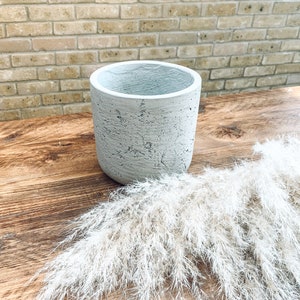 Puede incluir: Un macetero de hormigón gris con una superficie rugosa y texturizada. El macetero está vacío y se encuentra sobre una mesa de madera con un ramo de hierba de la pampa seca.