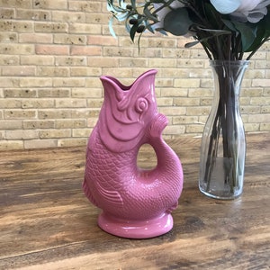 May include: A pink ceramic fish-shaped jug with a textured surface. The jug is on a wooden table.