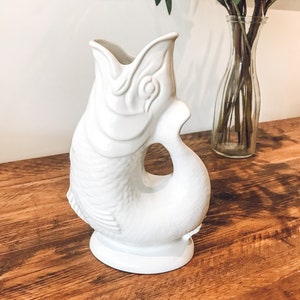 May include: White ceramic pitcher shaped like a fish. The pitcher has a wide mouth and a handle that is formed by the fish's tail. The pitcher is sitting on a wooden table.