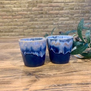 Op de afbeelding: Twee blauwe keramische mokken met een wit glazuurontwerp. De mokken staan op een houten oppervlak.