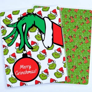May include: A rectangular planner cover featuring a white background with a repeating pattern of green Grinch faces wearing red Santa hats. A large green hand in a red sleeve holds a red ornament with the text Merry Grinchmas! in white.