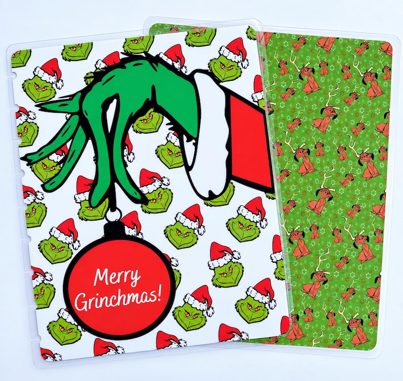 May include: A rectangular planner cover featuring a white background with a repeating pattern of green Grinch faces wearing red Santa hats. A large green hand in a red sleeve holds a red ornament with the text Merry Grinchmas! in white.