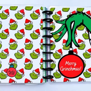 May include: A disc-bound planner featuring a repeating pattern of green Grinch faces wearing red Santa hats. The right page displays a large green hand holding a red Christmas ornament with the text Merry Grinchmas! in white script lettering.