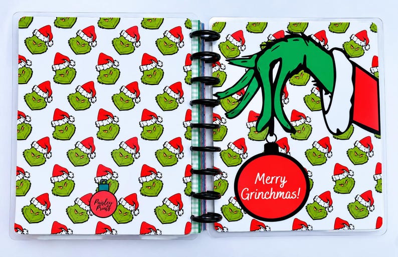 May include: A disc-bound planner featuring a repeating pattern of green Grinch faces wearing red Santa hats. The right page displays a large green hand holding a red Christmas ornament with the text Merry Grinchmas! in white script lettering.