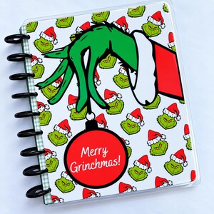 May include: A disc-bound notebook featuring a repeating pattern of green Grinch faces wearing red Santa hats. A large green hand in a red sleeve holds a red Christmas ornament with the white text Merry Grinchmas! printed on the front.