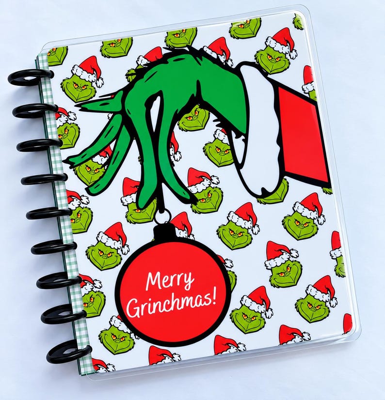 May include: A disc-bound notebook featuring a repeating pattern of green Grinch faces wearing red Santa hats. A large green hand in a red sleeve holds a red Christmas ornament with the white text Merry Grinchmas! printed on the front.