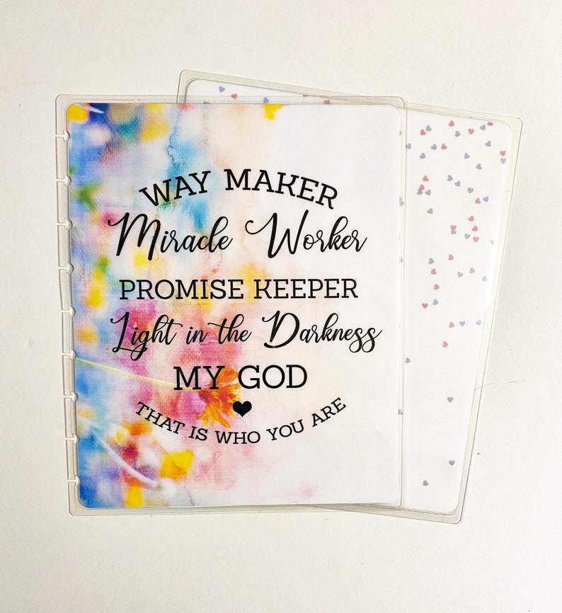 May include: Clear plastic planner cover with the text "WAY MAKER Miracle Worker PROMISE KEEPER Light in the Darkness MY GOD THAT IS WHO YOU ARE" over a watercolor floral background. A second cover with a heart pattern is visible.