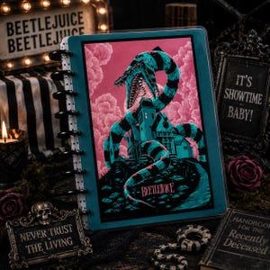 Könnte beinhalten: Ein scheibengebundenes Notizbuch mit einem türkis-schwarz gestreiften Sandwurm, der sich vor einem rosafarbenen, bewölkten Himmel um ein Haus windet. Auf dem Einband steht das Wort Beetlejuice in stilisierter Schrift. Das Notizbuch hat einen weißen Rücken mit schwarzen Scheiben.