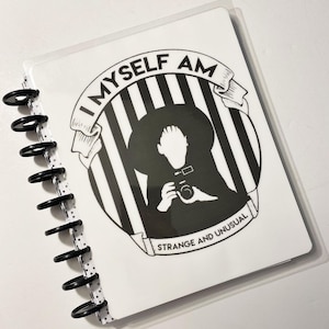 Puede incluir: Una cubierta de cuaderno en blanco y negro con un gráfico de una persona sosteniendo una cámara. El gráfico está rodeado de rayas blancas y negras y una pancarta que dice "Yo mismo soy extraño e inusual".