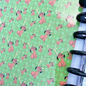 May include: A green notebook cover featuring a repeating pattern of a cartoon dog with antlers, interspersed with small white and red snowflake-like shapes. The notebook is bound with black plastic discs along the right side edge.