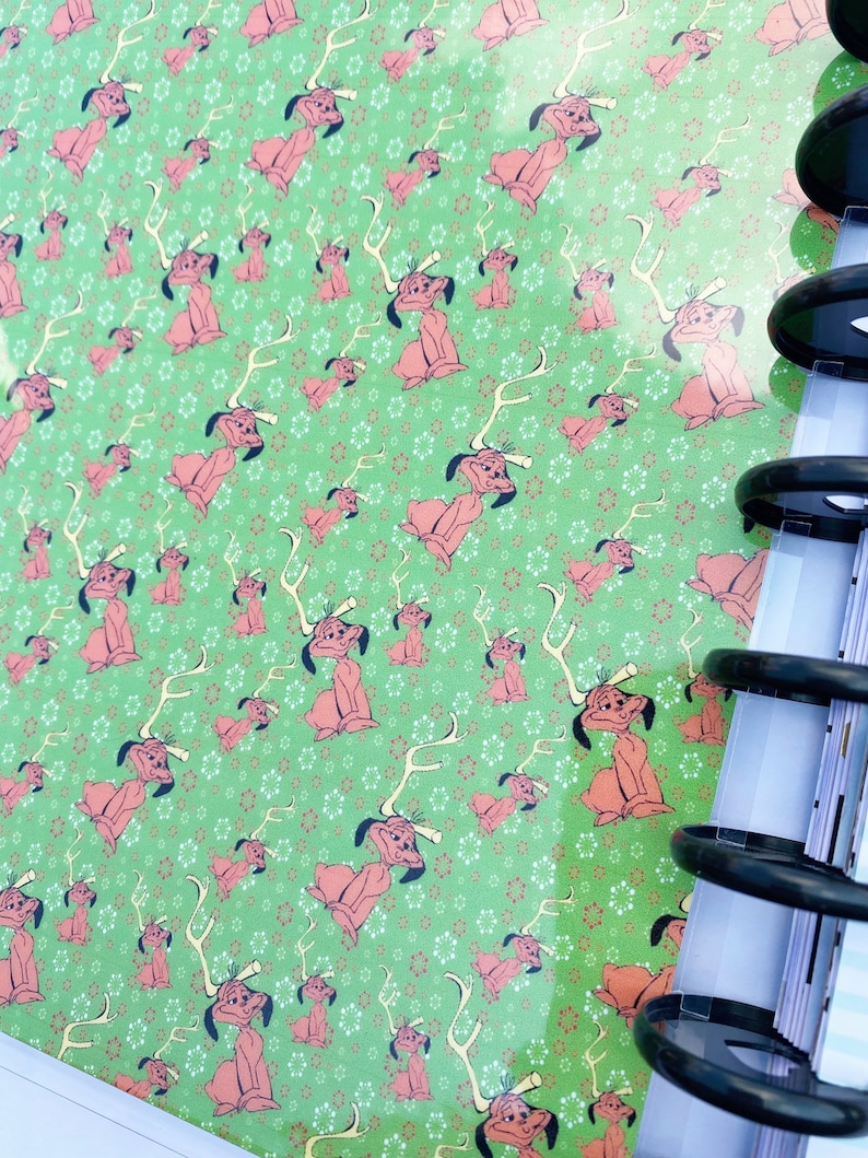May include: A green notebook cover featuring a repeating pattern of a cartoon dog with antlers, interspersed with small white and red snowflake-like shapes. The notebook is bound with black plastic discs along the right side edge.