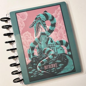 Puede incluir: Una agenda azul verdosa con una cubierta de plástico transparente que presenta una ilustración en blanco y negro de una criatura rayada con una casa en la espalda. La criatura está sobre un fondo rosa y verde con el texto "BEETLEJUICE" en blanco.