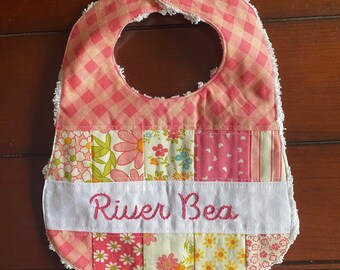 May include: A pink and white patchwork baby bib with a white trim and a yellow button. The bib is embroidered with the name "River Bea".