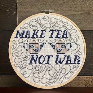 Pode incluir: Uma bordado de ponto cruz circular num bastidor de madeira apresenta a frase "MAKE TEA NOT WAR" em fio azul marinho. Duas chaleiras são bordadas sob as palavras. O desenho é rodeado por redemoinhos cinzentos sobre um fundo branco.