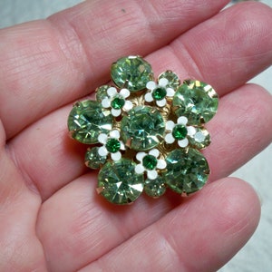 May include: A green and white rhinestone brooch with a floral design. The brooch is made of metal and has a gold finish.