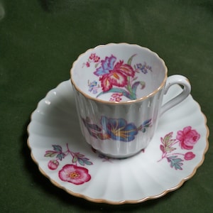 May include: A white teacup and saucer with a floral design. The cup has a gold rim and the saucer has a scalloped edge.