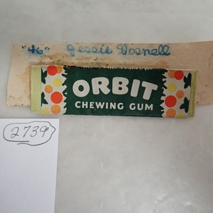 May include: A vintage pack of Orbit chewing gum with a green and white design, featuring the brand name in large white letters. The packaging includes a decorative pattern of orange and yellow circles. A small piece of paper with the name "Jessie Dosnell" is above the gum.