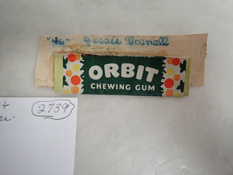 May include: A vintage pack of Orbit chewing gum with a green and white design, featuring the brand name in large white letters. The packaging includes a decorative pattern of orange and yellow circles. A small piece of paper with the name "Jessie Dosnell" is above the gum.