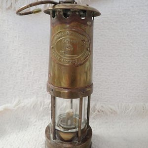 May include: Antique brass miner's lamp with a glass enclosure. The lamp has a handle and an oval plaque with text that reads "S. Thomas & Williams Ltd. Cambrian, No. Maker - Aberdare, Wales."