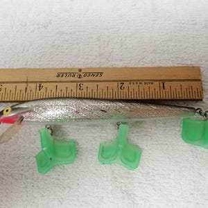 May include: A silver fishing lure with a pink and yellow accent, and three green plastic fins. A wooden ruler with black markings is placed next to the lure. The ruler is marked in inches.