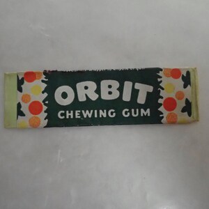 May include: A rectangular package of Orbit chewing gum. The wrapper is dark green with the word "ORBIT CHEWING GUM" in white. The ends of the wrapper have a pattern of orange, yellow, and white circles.