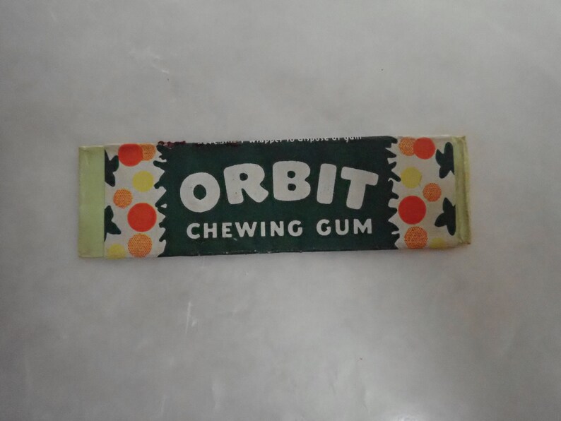 May include: A rectangular package of Orbit chewing gum. The wrapper is dark green with the word "ORBIT CHEWING GUM" in white. The ends of the wrapper have a pattern of orange, yellow, and white circles.