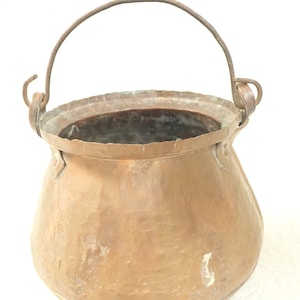 May include: A vintage copper pot with a wire handle. The pot has a rounded bottom and a wide opening. The copper has a patina and shows signs of age.