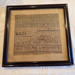 May include: A framed sampler with black embroidery on a beige background. The sampler includes the alphabet, numbers, and the text "WXYZ in the Year of our Lord 1830 mar x am 8 years T".