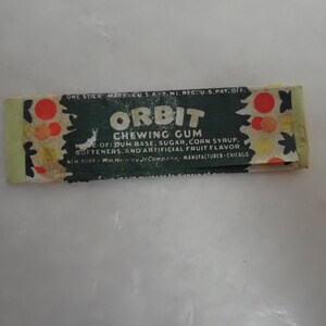 May include: Vintage Orbit chewing gum wrapper. The wrapper is rectangular, with a dark green background and the word "ORBIT" in large white letters. The wrapper also includes the text "CHEWING GUM" and the ingredients. The wrapper has a light green border and red and orange circular designs.