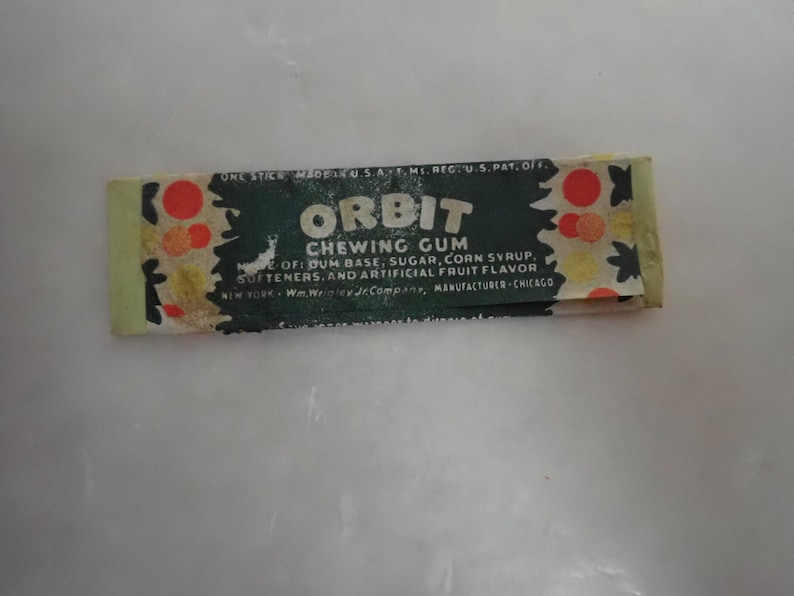 May include: Vintage Orbit chewing gum wrapper. The wrapper is rectangular, with a dark green background and the word "ORBIT" in large white letters. The wrapper also includes the text "CHEWING GUM" and the ingredients. The wrapper has a light green border and red and orange circular designs.