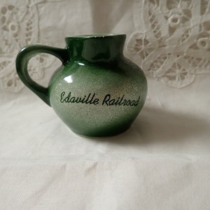 May include: A small, green ceramic pitcher with a handle. The pitcher has a rounded body and a speckled, silver-toned finish on the front. The words "Edaville Railroad" are written in black script on the front.