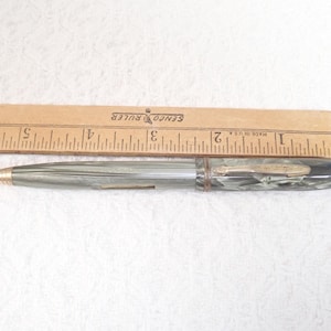 May include: A vintage mechanical pencil with a gold-colored tip and a gray and black marbled body. A wooden ruler with inch markings is placed next to the pencil. The ruler is approximately 6 inches long.