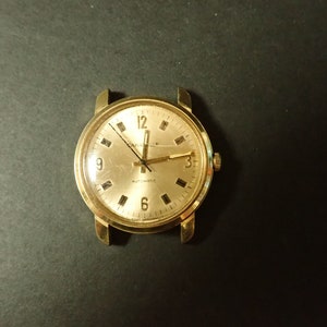 May include: A gold-tone wristwatch with a round face and black hands. The watch face has black Roman numeral hour markers and the words "Caravelle Automatic" printed on it.