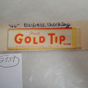 May include: A vintage Fruit Gold Tip Gum wrapper with a yellow background and red lettering. The wrapper has a handwritten inscription that reads "'46' Barbara Shockley".