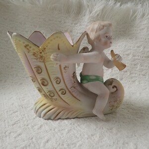 May include: A porcelain figurine of a cherub playing a horn, sitting on a yellow and pink shell-shaped planter.