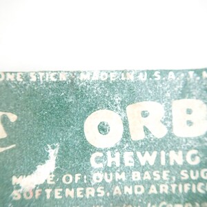 May include: Close-up of a vintage Orbit chewing gum package. The package features the word "ORBIT" in large, bold letters, with the words "CHEWING GUM" below. The package also includes text about the ingredients and the phrase "MADE IN U.S.A."