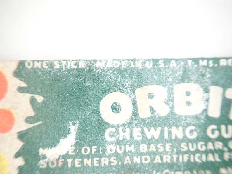 May include: Close-up of a vintage Orbit chewing gum package. The package features the word "ORBIT" in large, bold letters, with the words "CHEWING GUM" below. The package also includes text about the ingredients and the phrase "MADE IN U.S.A."