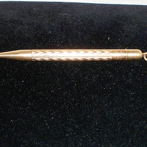 May include: A gold-toned mechanical pencil with a black and white patterned barrel and a ring at the end. The pencil is lying on a black velvet surface.