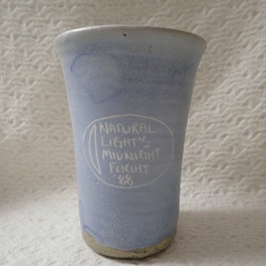 May include: A light blue ceramic mug with a white inscription that reads "NATURAL LIGHT'S MIDNIGHT FLIGHT '88".