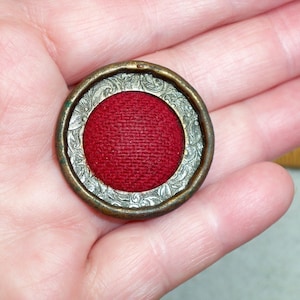 May include: A round, silver-colored pin with a red fabric center. The pin has a decorative, floral design around the edge.