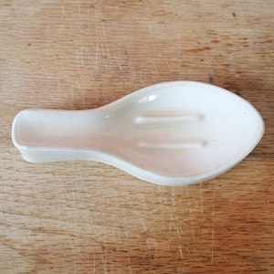 May include: White ceramic spoon rest with a slightly curved shape and two ridges on the inside.