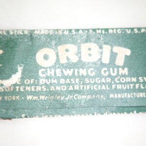 May include: Close-up of a vintage Orbit chewing gum wrapper. The wrapper is green with white text that reads "Orbit Chewing Gum." Ingredients include gum base, sugar, corn syrup, and artificial fruit flavor. Made in the U.S.A.