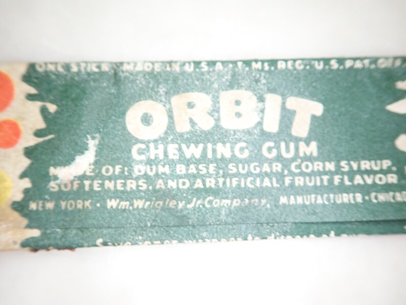 May include: Close-up of a vintage Orbit chewing gum wrapper. The wrapper is green with white text that reads "Orbit Chewing Gum." Ingredients include gum base, sugar, corn syrup, and artificial fruit flavor. Made in the U.S.A.