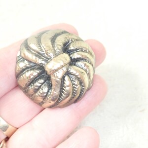 May include: A silver-toned metal button with a knotted design. The button is round and has a raised, textured surface.